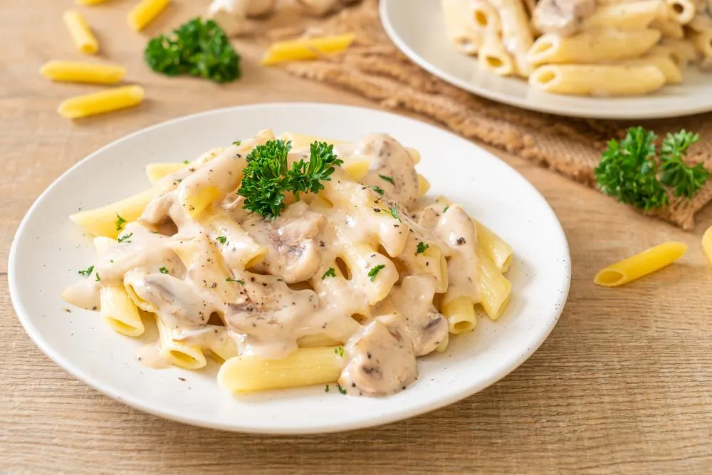 Prato de macarrão penne com molho de creme de cogumelos, decorado com salsa fresca, servido em prato branco sobre mesa de madeira.