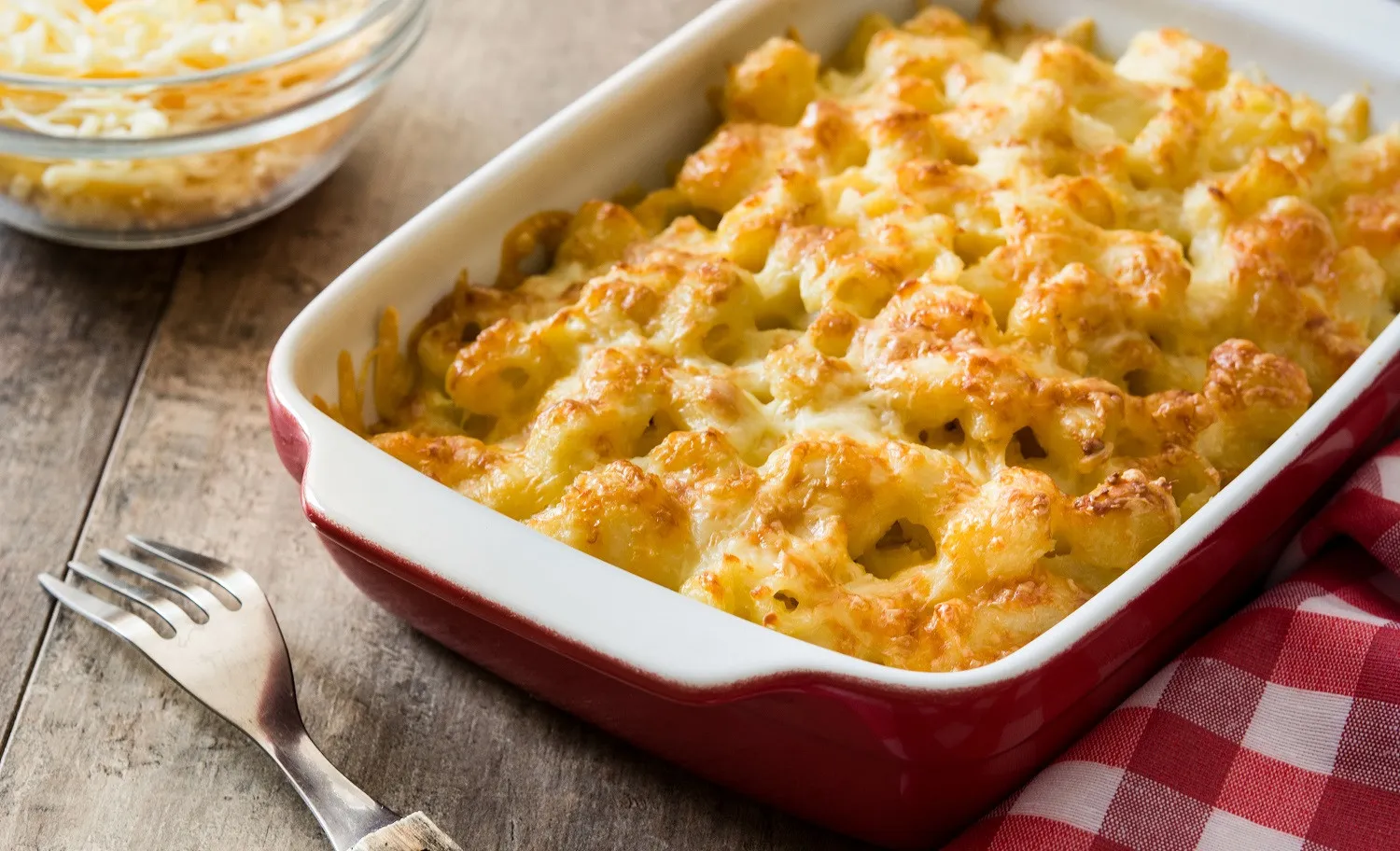Imagem de uma travessa de macarrão de forno com queijo derretido e crosta dourada, servido em uma mesa de madeira com uma colher ao lado.