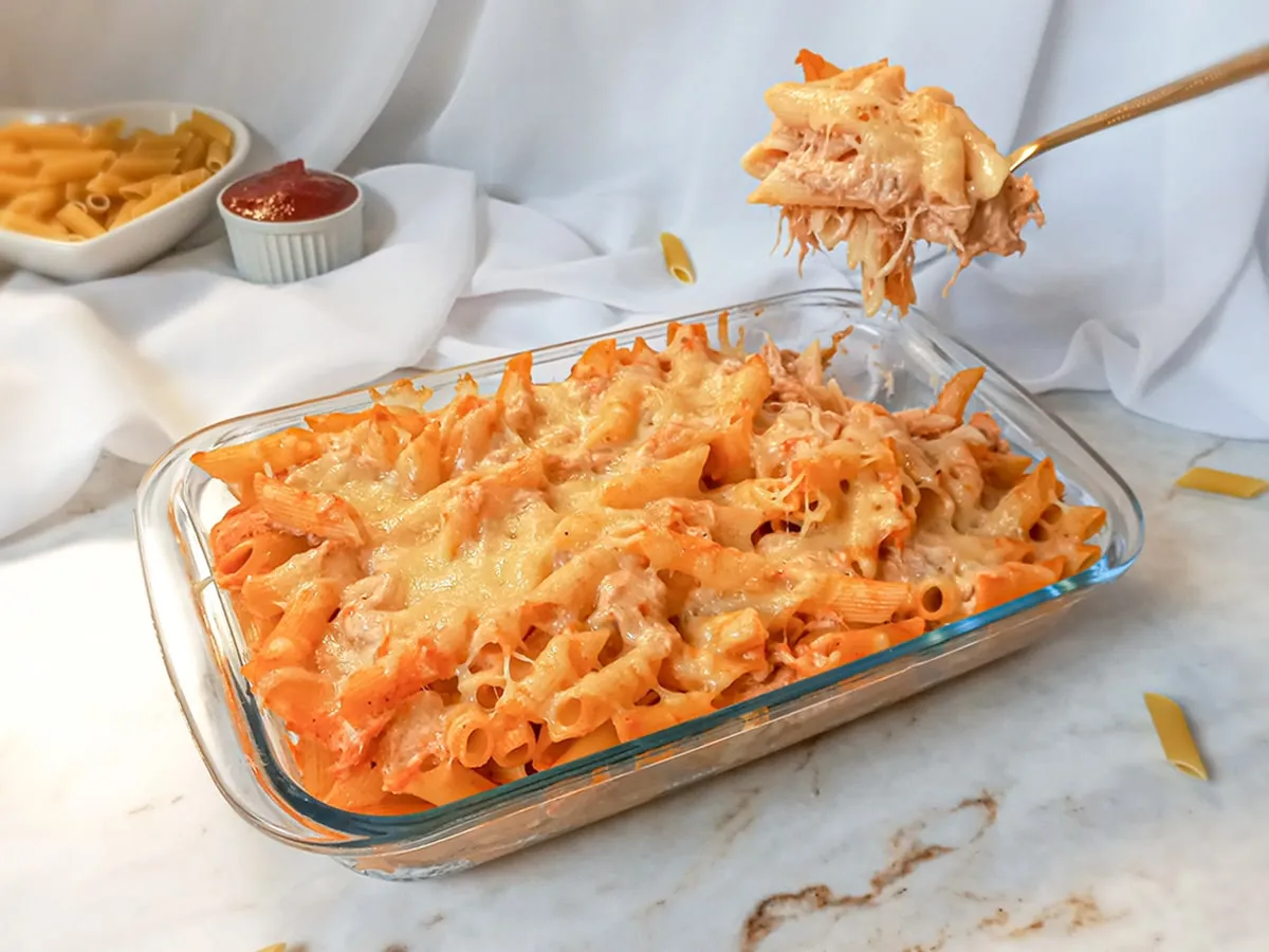 Prato de macarrão com molho de queijo e tomate em uma travessa de vidro, ao lado de molho de tomate e queijo em tigelas, com uma colher servindo uma porção