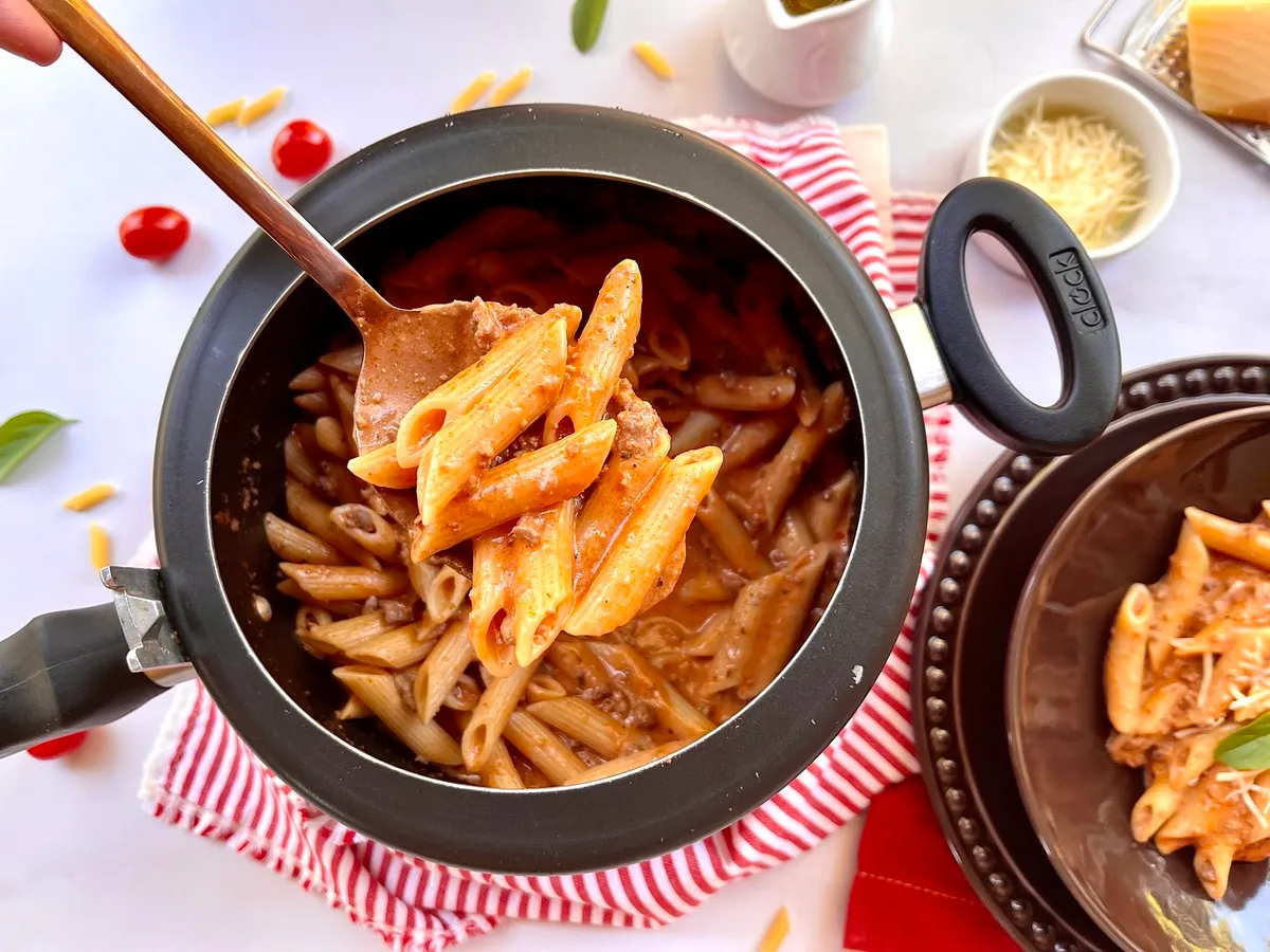 Imagem de uma panela de macarrão penne com molho, junto de ingredientes como queijo ralado e tomates cereja, ideal para uma receita de macarrão caseiro