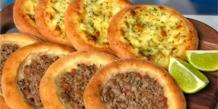 Duas opções de esfiha com queijo e carne bovina, acompanhadas de fatias de limão, em uma bandeja de madeira.