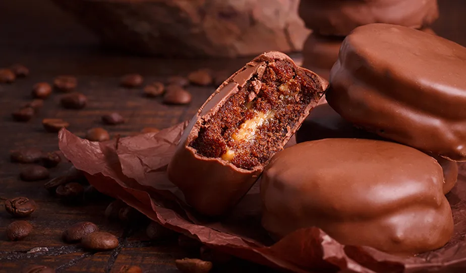 Imagem de docinhos de chocolate recheados, com cobertura de chocolate ao leite, sobre uma superfície de madeira, decorados com grãos de café ao redor.