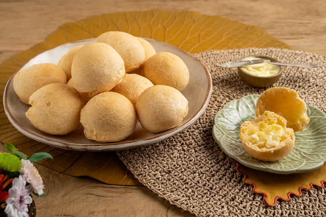 Deliciosos pão de queijo mineiro tradicional feito com queijo artesanal, servidos em um prato de cerâmica, acompanhado de manteiga amanteigada e decorado com folhas secas.