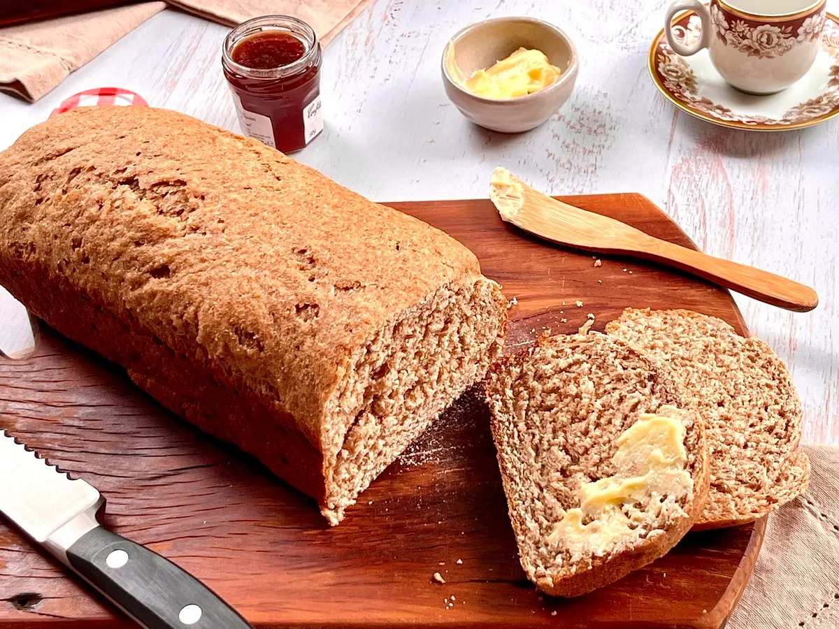 Pão de trigo caseiro sendo cortado com manteiga e geleia na tábua de madeira, ideal para café da manhã ou lanche.