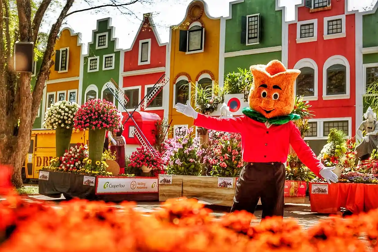 Personagem de personagem de fantasia com cabeça de animal, vestido de vermelho, em feira com flores coloridas e casas decorativas ao fundo, em um ambiente festivo para o dia das crianças.