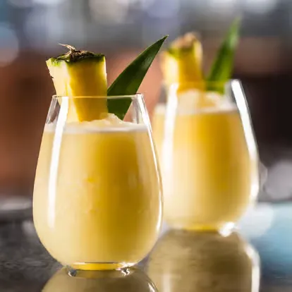 Dois copos de drinque de cor amarelo com fatia de abacaxi e folha de hortelã decorando a bebida, em fundo desfocado. Bebida refrescante e deliciosa.