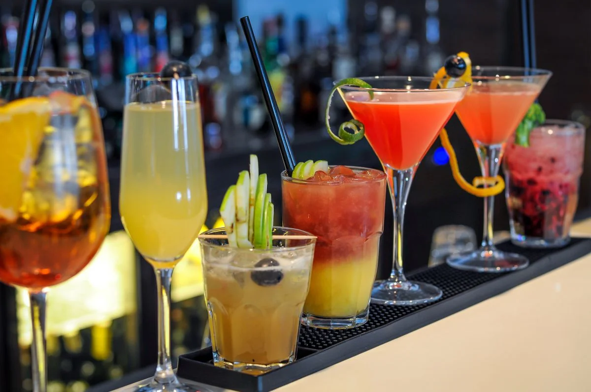 Diferentes tipos de coquetéis coloridos e decorados, dispostos em um balcão de bar, prontos para serem servidos em uma festa ou happy hour.