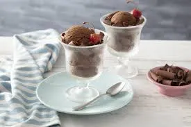 Sorvetes de chocolate em copos de vidro decorados com frutas vermelhas, servidos em uma mesa moderna com colher e toalha de tecido azul claro.