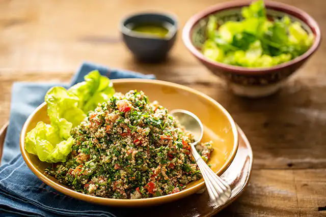 Salada de tabule com legumes frescos em uma tigela de cerâmica, acompanhada de folhas verdes e molho em uma mesa de madeira, perfeita para uma refeição saudável.