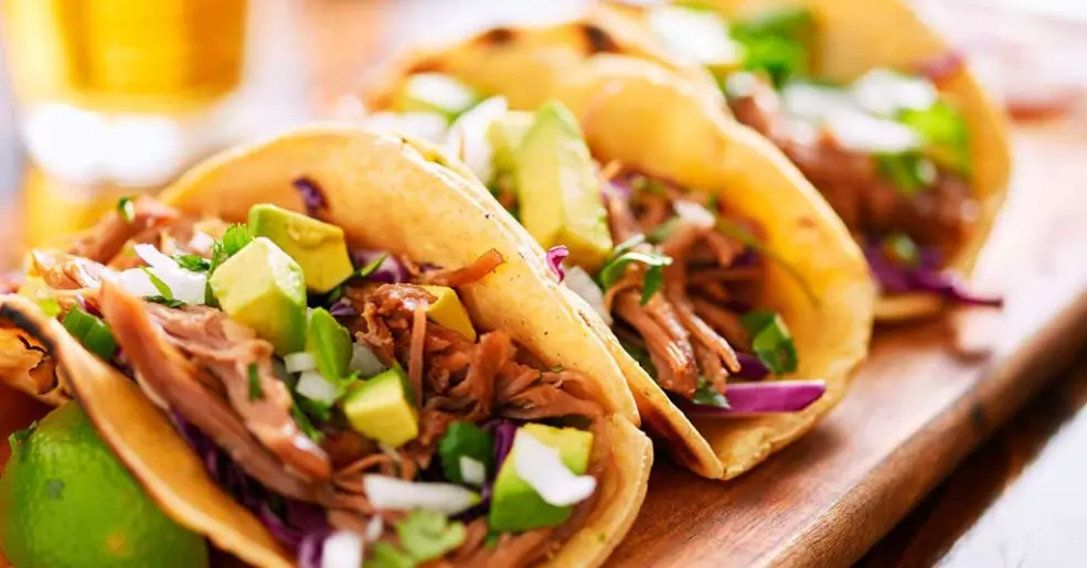 Três tacos de carne com guacamole, repolho roxo, cebola e verde em uma tábua de madeira, ideal para apreciar uma refeição mexicana deliciosa.