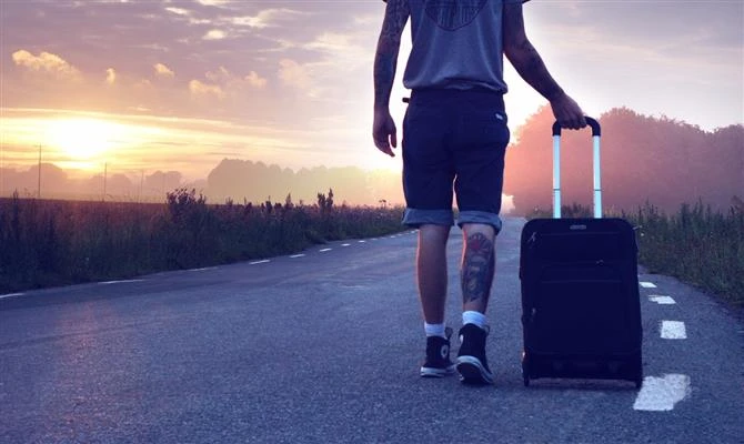 Pessoa caminhando na estrada com uma mala de viagem ao pôr do sol, simbolizando viagem e aventura em busca de novas experiências.