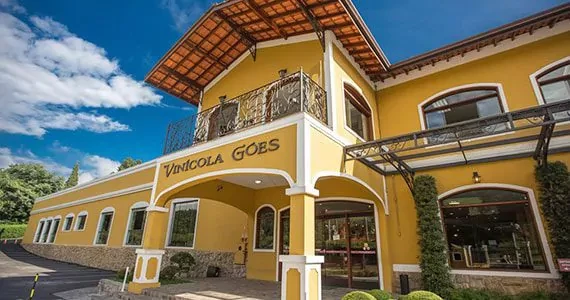 Imagem de uma charmosa casa de arquiteta italiana Vinícola Goes, com fachada amarela, varanda com ferro trabalhado e grande área de lazer ao ar livre, sob céu azul.