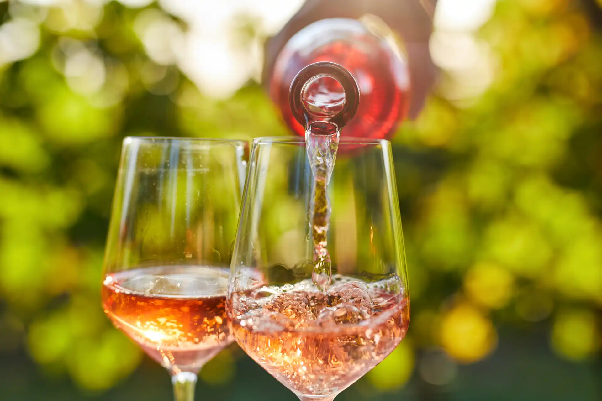 Vinho rosé sendo servido em taças em ambiente ao ar livre com fundo desfocado de folhas verdes, perfeito para momentos de celebração ao ar livre.