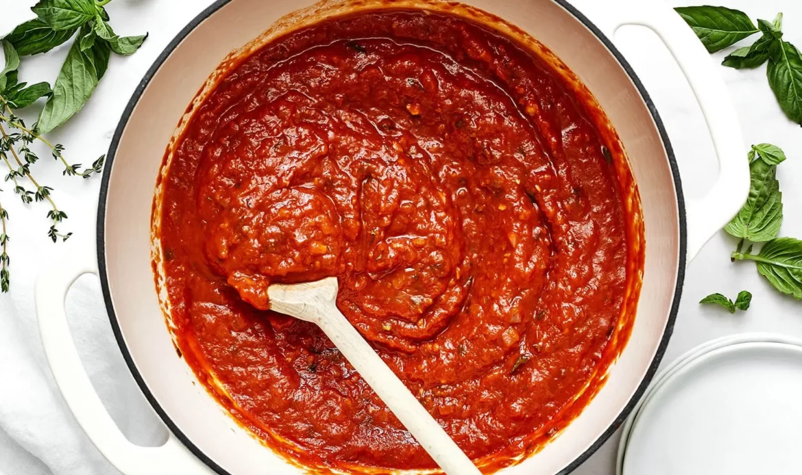 Molho marinara com textura cremosa em panela, rodeado de ervas aromáticas como manjericão e orégano, ideal para massas e pizzas.