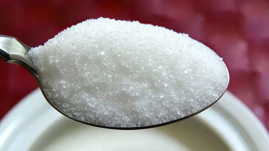 Imagem de adoçante em uma colher de metal, destaque para a textura granulada, com fundo levemente desfocado branco e vermelho, ideal para conteúdo sobre ingredientes de cozinha.