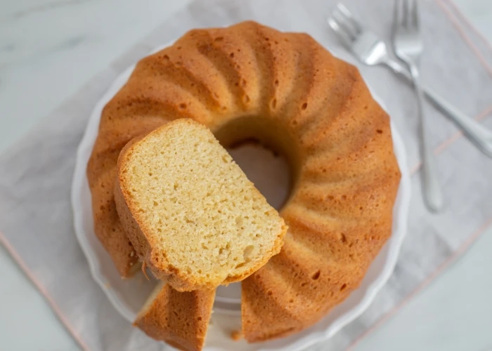 Bolo com pedaço cortado, apresentado em prato branco sobre mesa com utensílios ao fundo, perfeito para café da manhã ou lanche