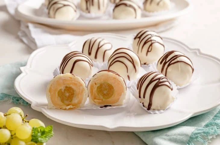 Imagem de trufas de chocolate branco recheadas com uva, algumas decoradas com riscas de chocolate, apresentadas em um prato decorativo branco para festa.