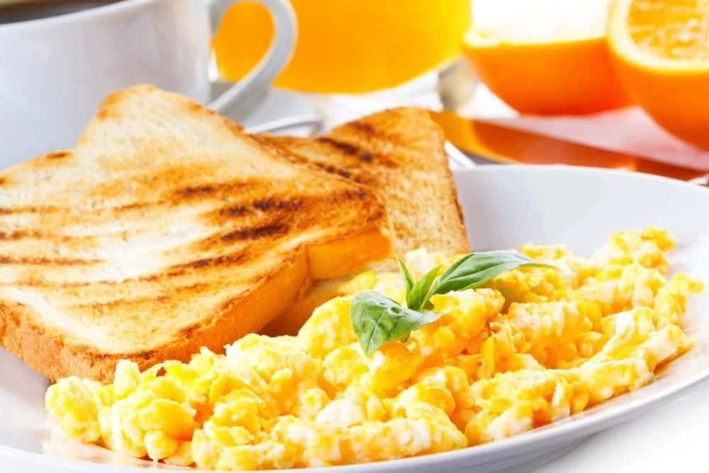 Imagem de café da manhã com ovos mexidos, torradas grelhadas, suco de laranja e uma fatia de laranja fatiada ao fundo, ideal para um café da manhã nutritivo.