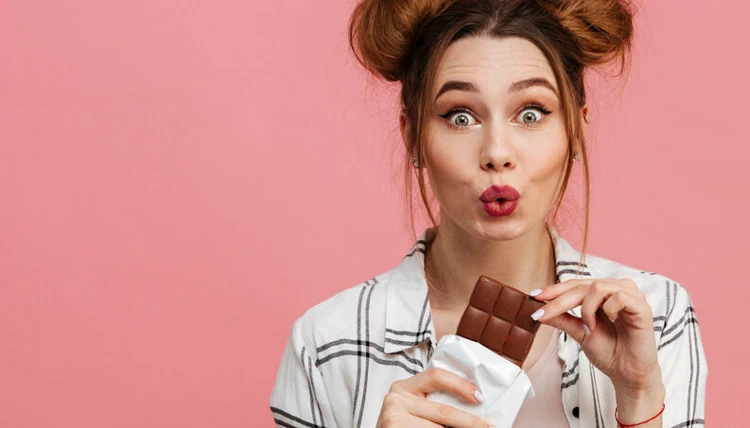 Jovem mulher com cabelo de coque feito em dois coques laterais, vestindo blusa xadrez, segurando uma barra de chocolate e fazendo expressão de surpresa, contra fundo rosa.