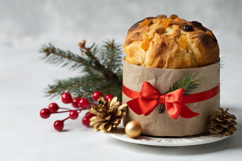 imagem de panetone de natal em cima de prato, enrolado em um papel e fita vermelha com laço. Atrás, ramas de natal.