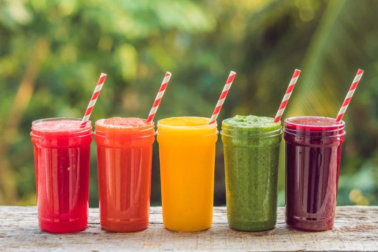 Cinco copos de sucos naturais de cores diferentes, incluindo vermelho, laranja, amarelo, verde e roxo, com canudos vermelhos e branco, dispostos em uma mesa de madeira ao ar livre, ideal para consumo de líquidos saudáveis e refrescantes.