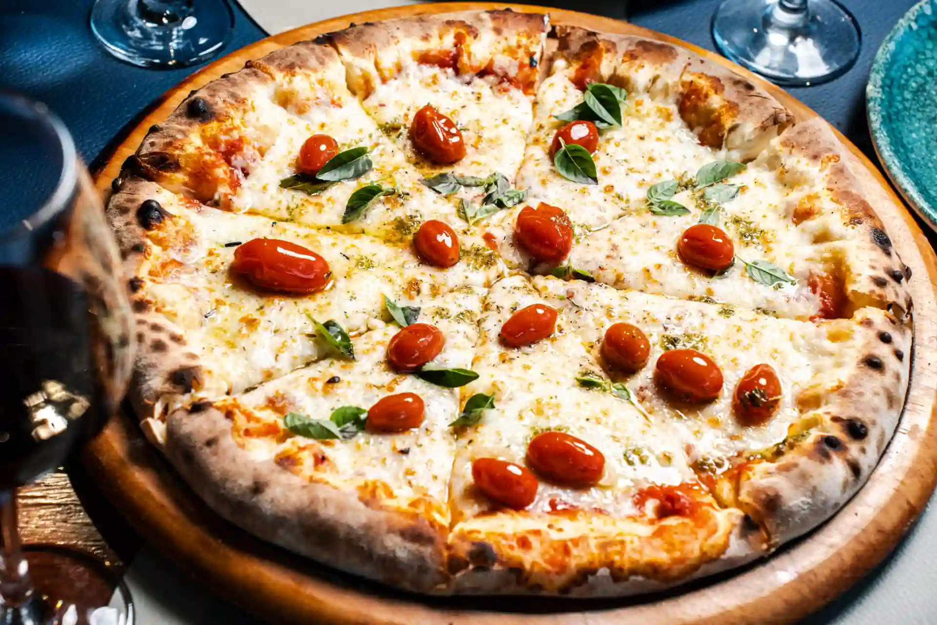 Pizza marguerita com tomate cereja, queijo derretido, manjericão e massa crocante, servida em uma mesa com taças de vinho ao fundo.