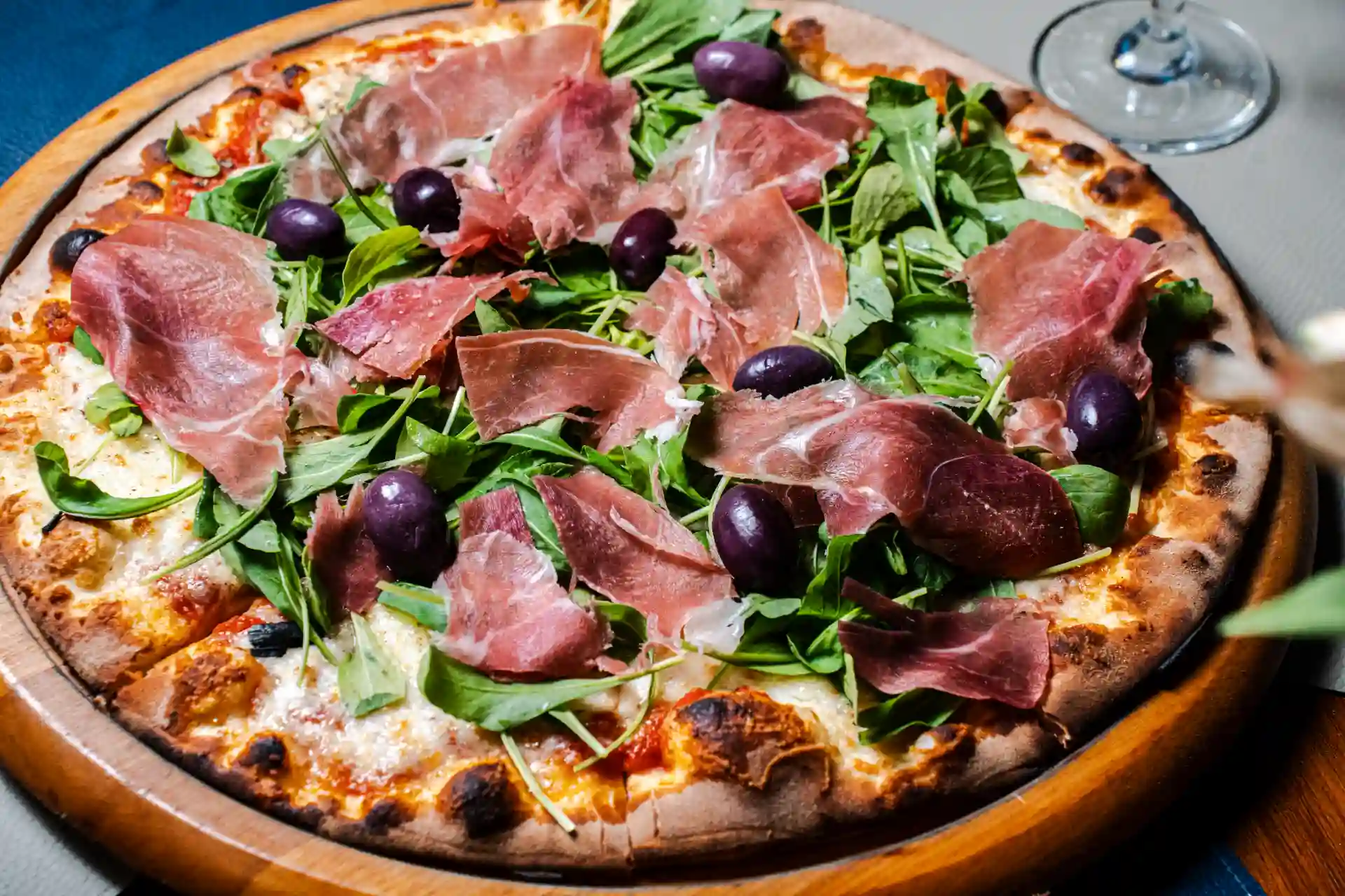 Pizza com presunto, rúcula, azeitonas pretas e queijo derretido em uma massa crocante, servido em uma pedra de pizza com vidro de vinho ao lado.