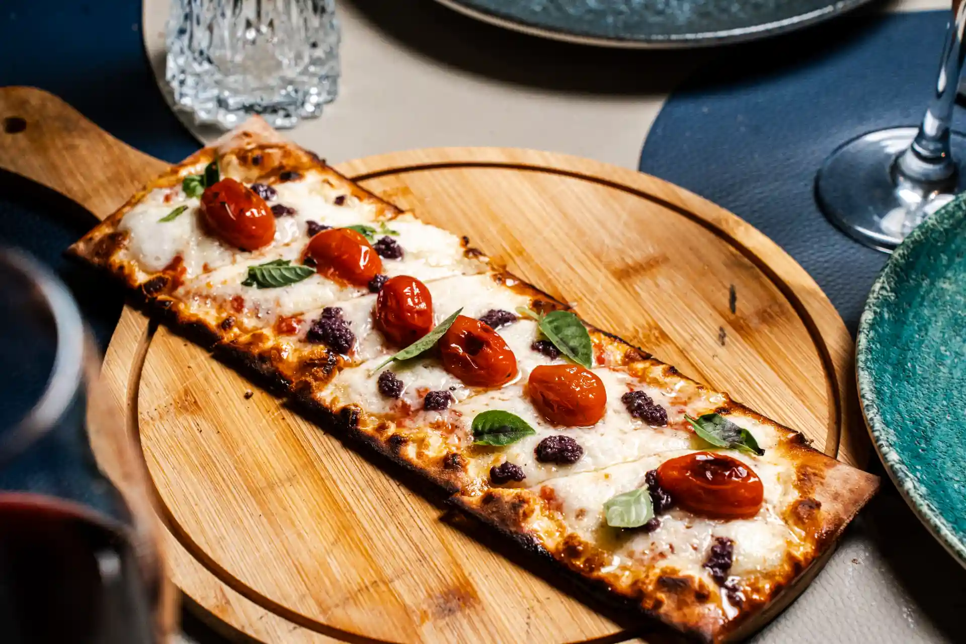 Pizza retangular com queijo, tomate cherry, folhas de manjericão e azeitonas, servida em uma tábua de madeira em ambiente de restaurante.