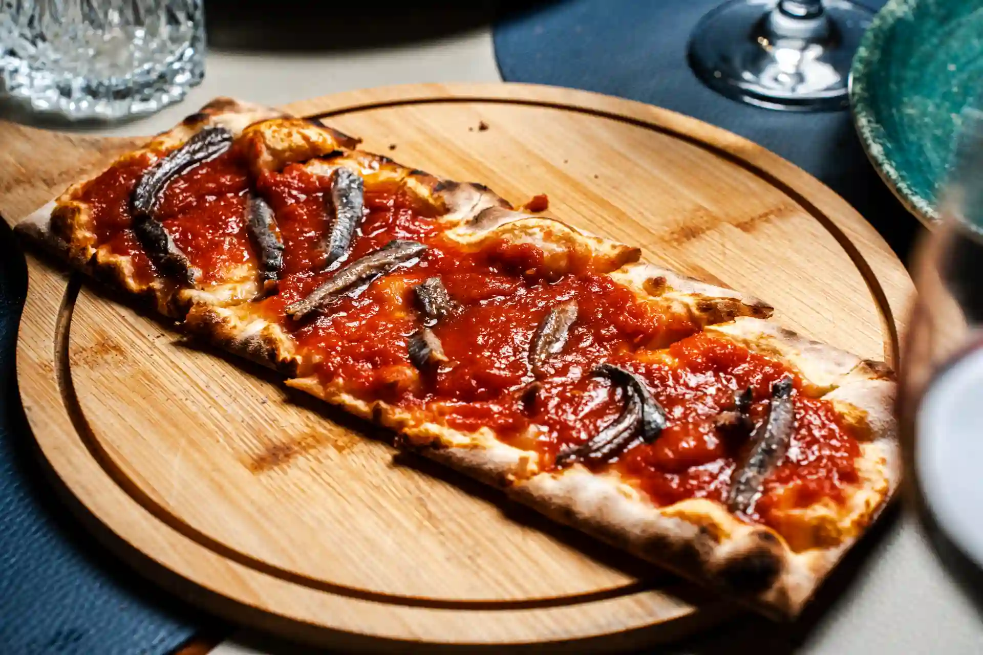 Pizza de alho com molho de tomate e sardinhas assadas em uma tábua de madeira, acompanhada de taças de vinho e utensílios de jantar.