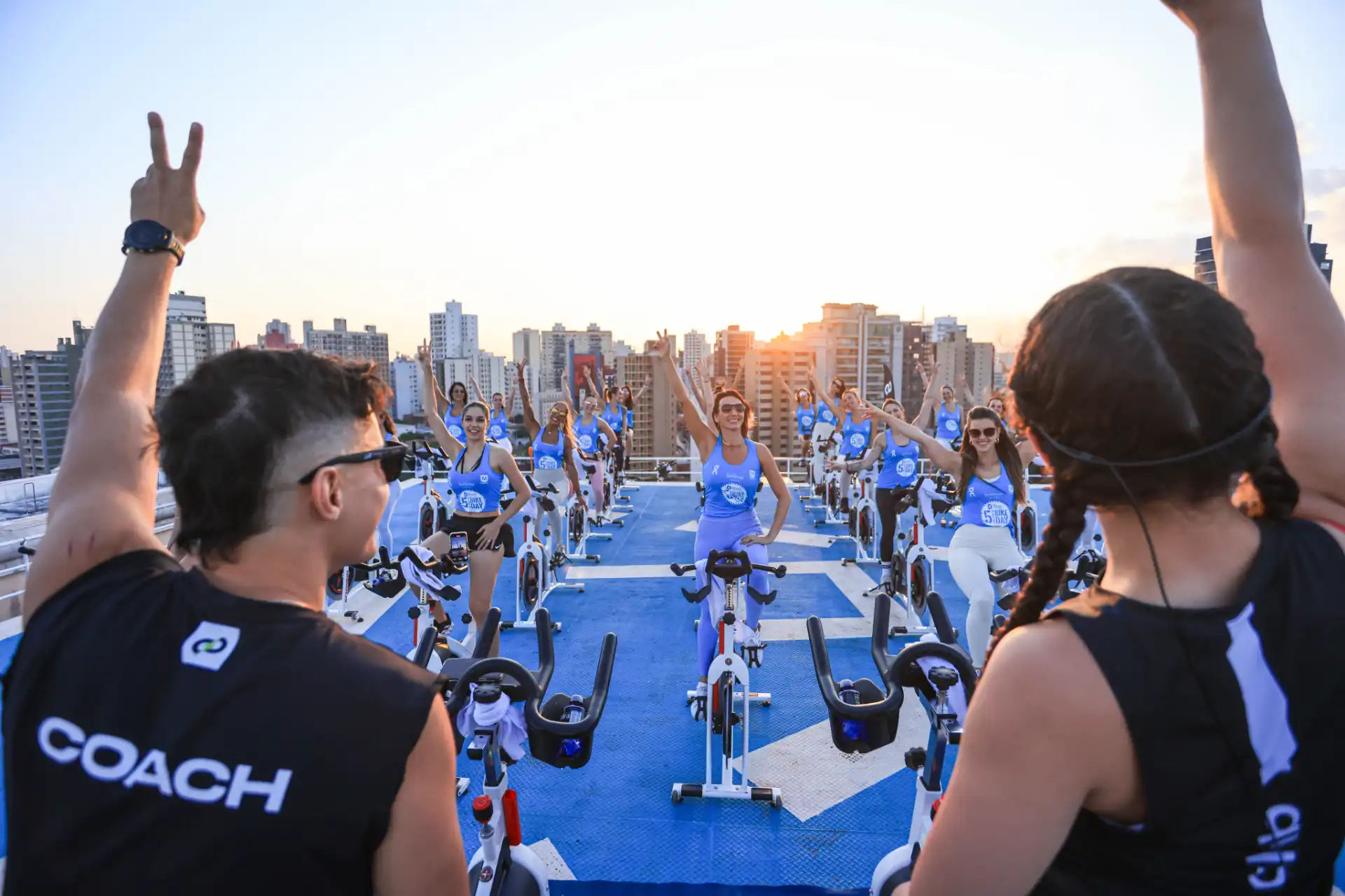 Grupo de pessoas participando de aula de spinning ao pôr do sol em rooftop na cidade, com instrutores e ciclistas animados.