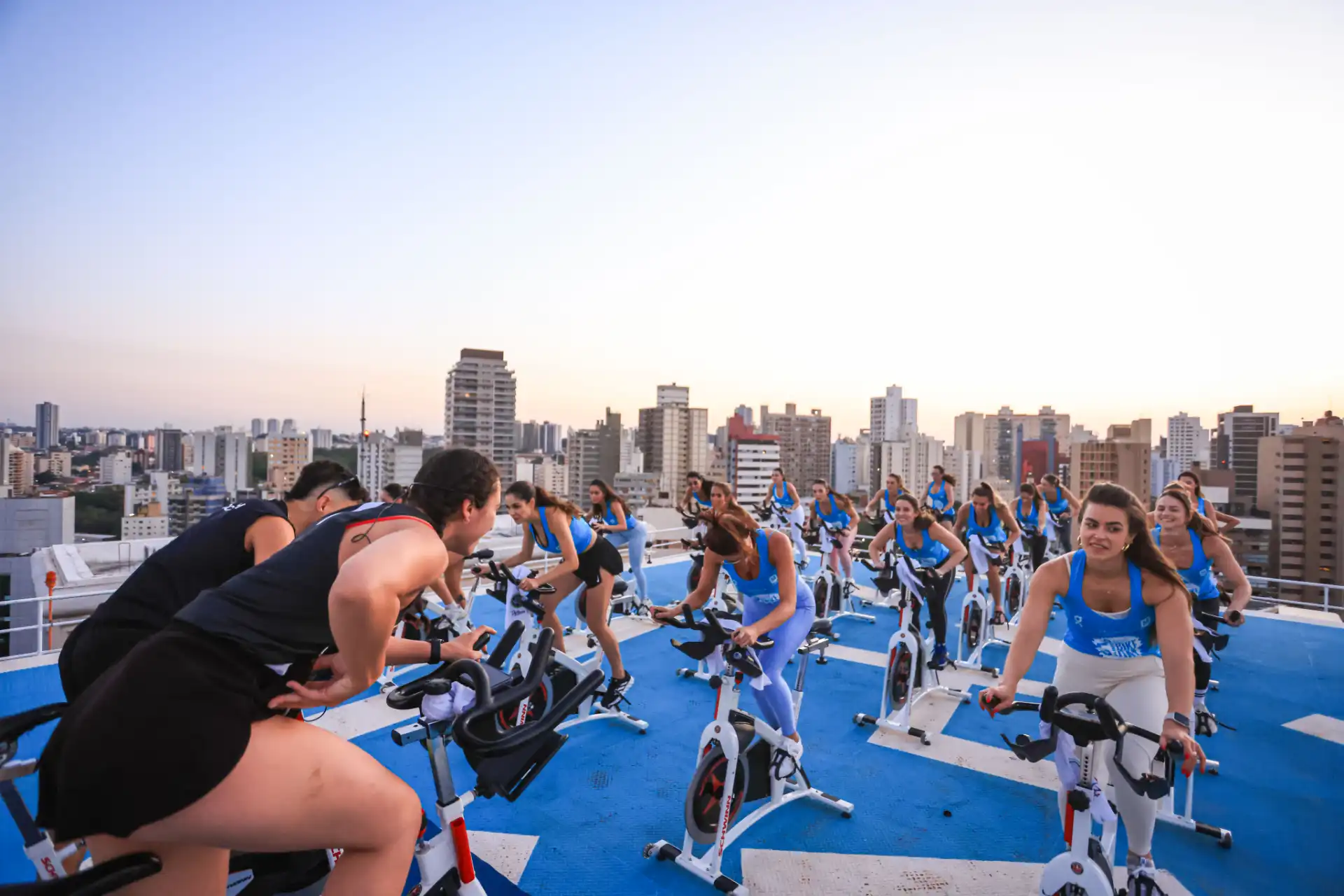 Grupo de pessoas participando de aula de spinning em um terraço com vista para a cidade ao entardecer, com várias bicicletas de spinning.