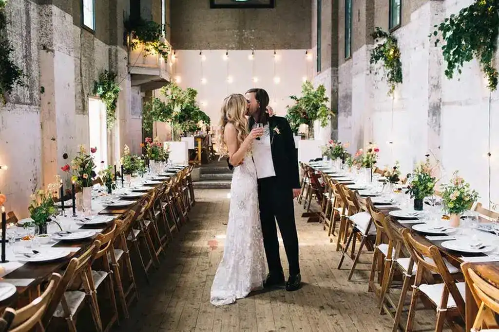Casal de noivos se beijando em cerimônia de casamento ao longo de uma longa mesa decorada com flores e velas, em ambiente rústico e elegante.