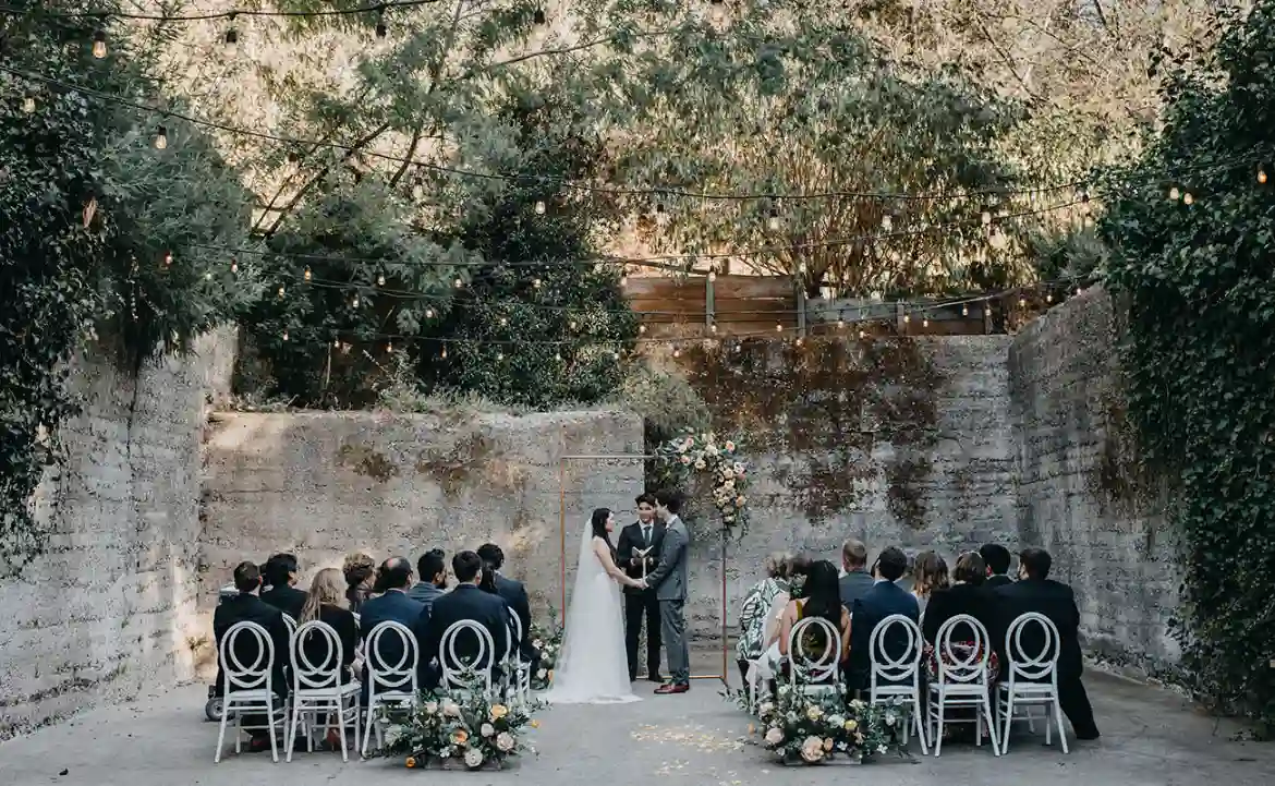 Cerimônia de casamento ao ar livre em um ambiente rústico, com convidados sentados, flores e árvores decorativas ao fundo.