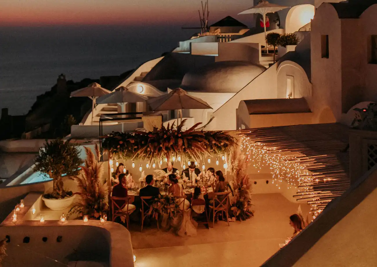 Celebração ao ar livre em um ambiente sofisticado com decoração de luzes, mesas elegantes e vista para o pôr do sol no topo de uma construção moderna.
