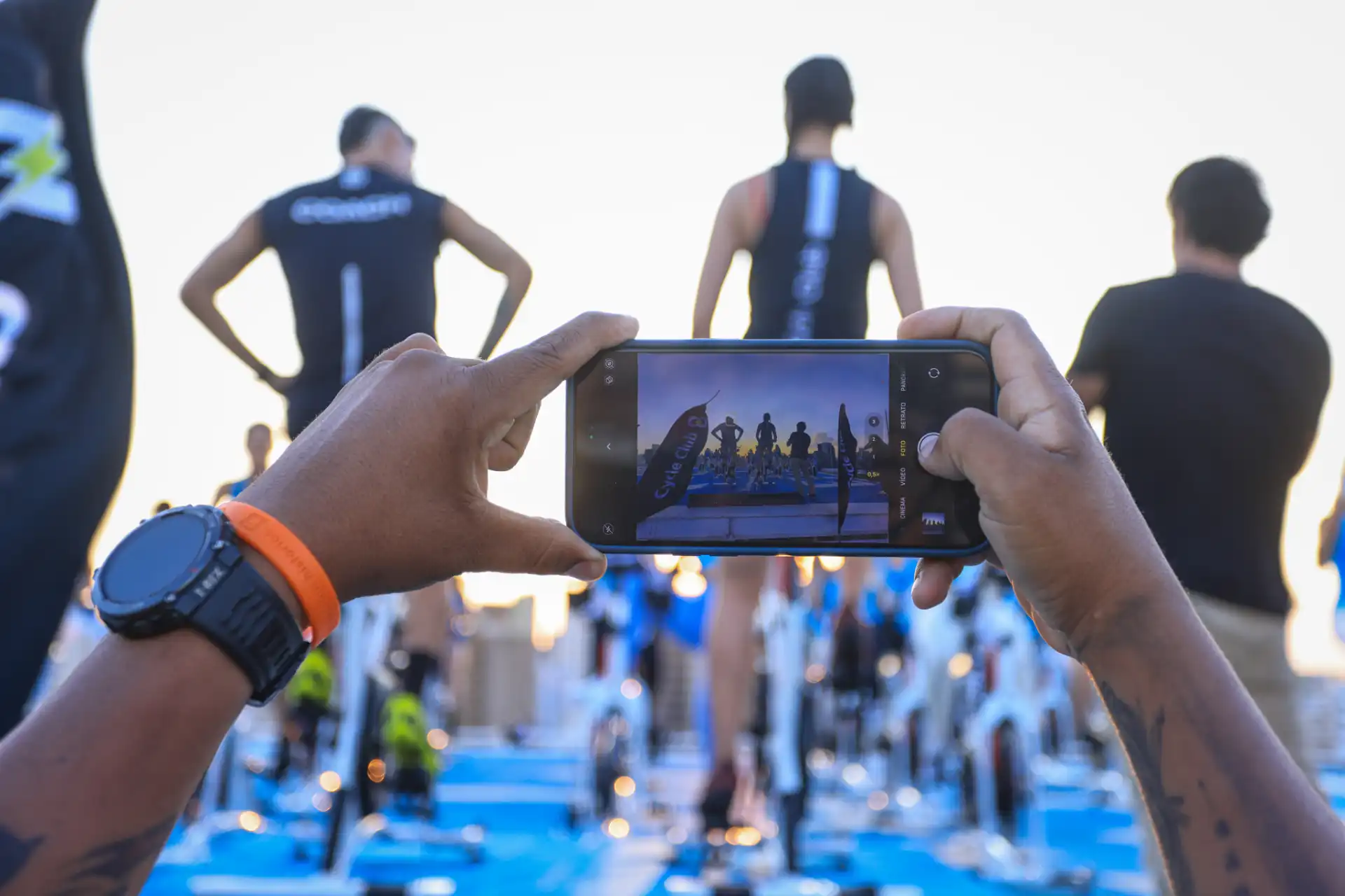 Pessoa registrando com o celular uma competição de corrida com atletas na linha de largada, ao ar livre com luz natural, foco na ação.