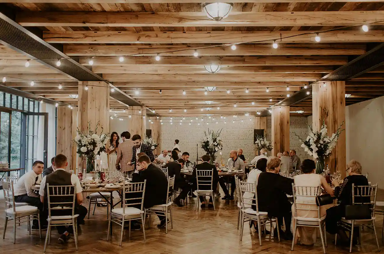 Casamento com convidados em ambiente elegante com decoração de flores e iluminação quente, no espaço de eventos com teto de madeira e estilo sofisticado.