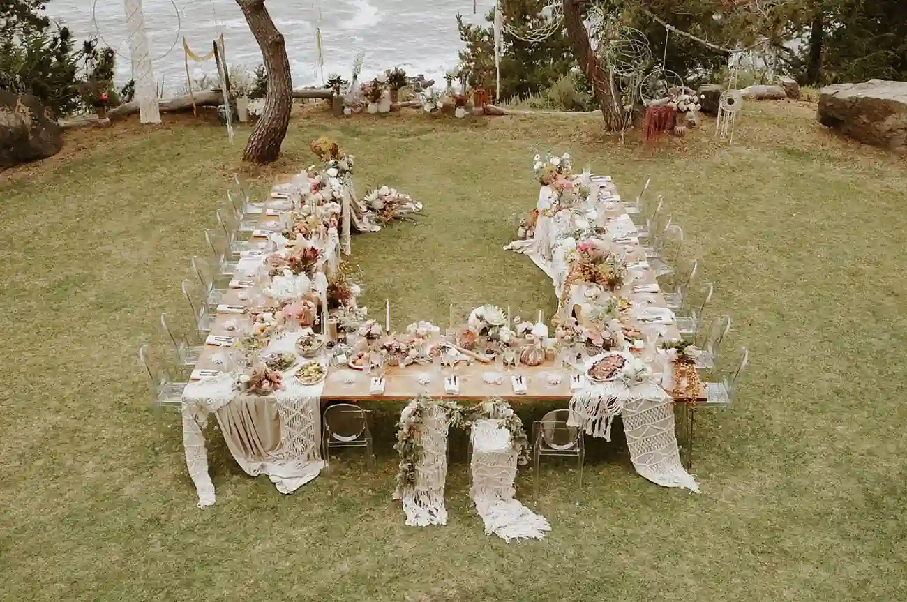 Mesa de festa de casamento ao ar livre decorada com flores, toalhas de renda e pratos finos, localizada em um espaço verde perto de uma área com água e árvores.