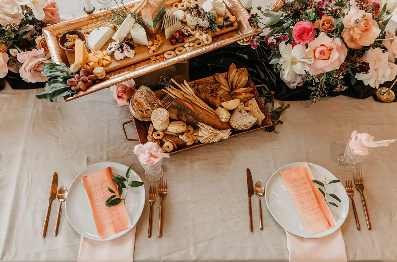 Mesa decorada com tábua de queijos, frutas secas e pães, rodeada por arranjos de flores em um cenário de festa elegante.