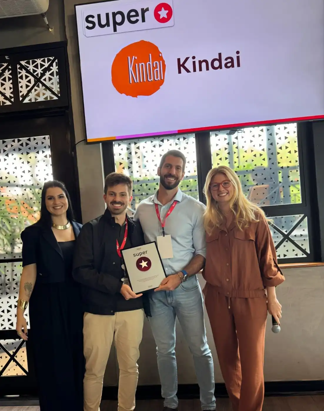 Grupo de pessoas sorrindo em uma cerimônia de premiação com banner do evento 'super Kindai' ao fundo e uma tela exibindo o mesmo logo.