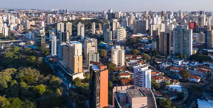 Imagem de uma vista aérea de uma cidade com muitos edifícios altos durante o dia, com áreas verdes visíveis, representando a urbanização moderna.