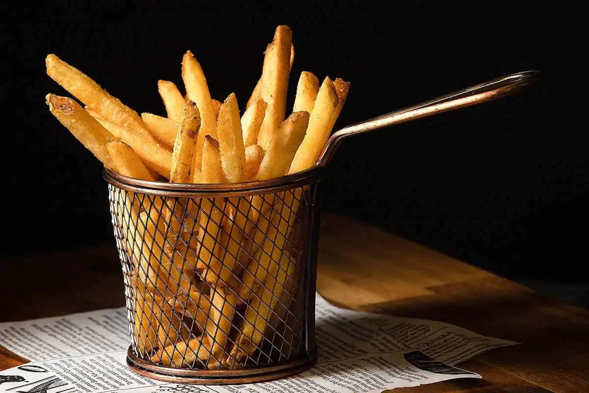 Porção de batatas fritas crocantes e douradas em cesto de metal sobre mesa de madeira, com fundo escuro destacando a comida.