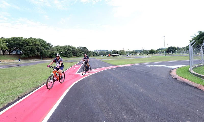 Ciclistas pedalando em ciclovia com pista de cor vermelha e preto, rodeados de natureza com árvores e céu claro, ideal para atividades ao ar livre.