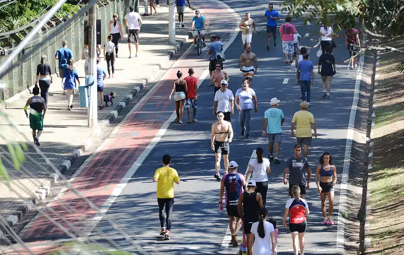 Imagem de pessoas caminhando, correndo e pedalando em uma ciclovia arborizada em um parque da cidade, com ambientes ao ar livre e espaço para atividades físicas ao ar livre.