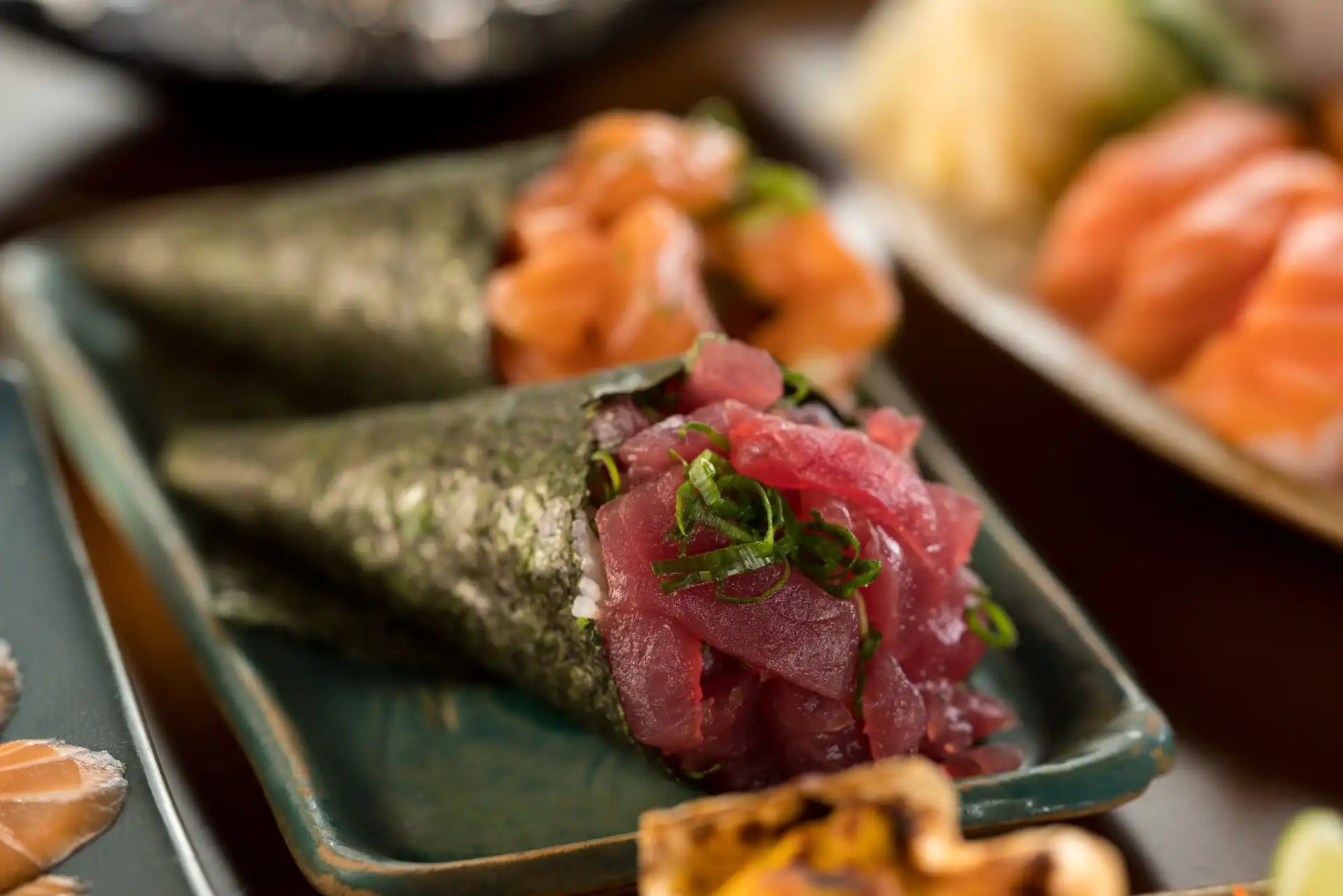 Prato de temaki de atum, envolvido por alga nori, servido em prato decorado, próximo a outras opções de sashimi de salmão e camarão.