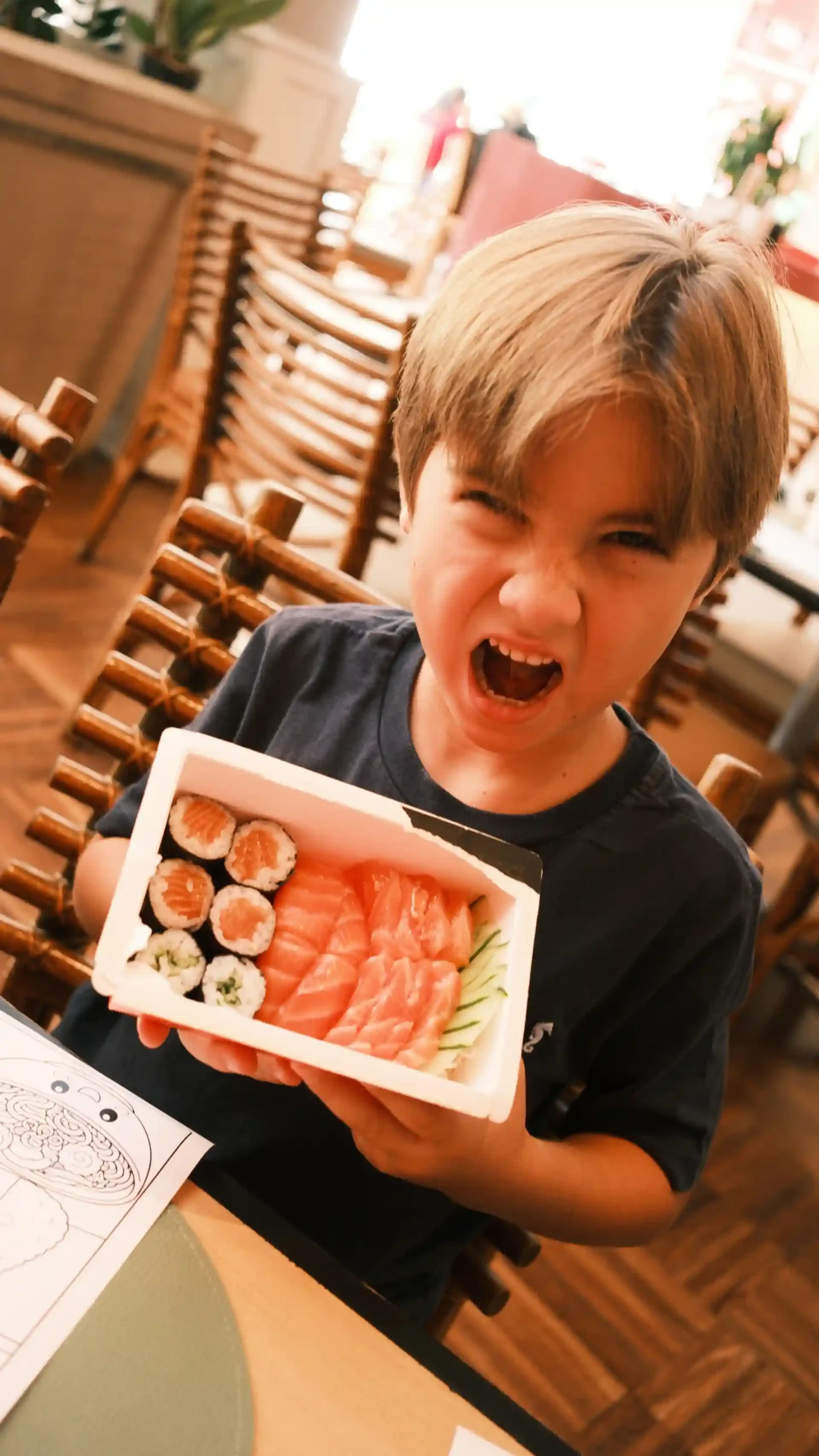 Criança feliz segurando uma caixa de sushi em restaurante, expressão de alegria, sushi variado, ambiente acolhedor, jantar entre amigos ou família.