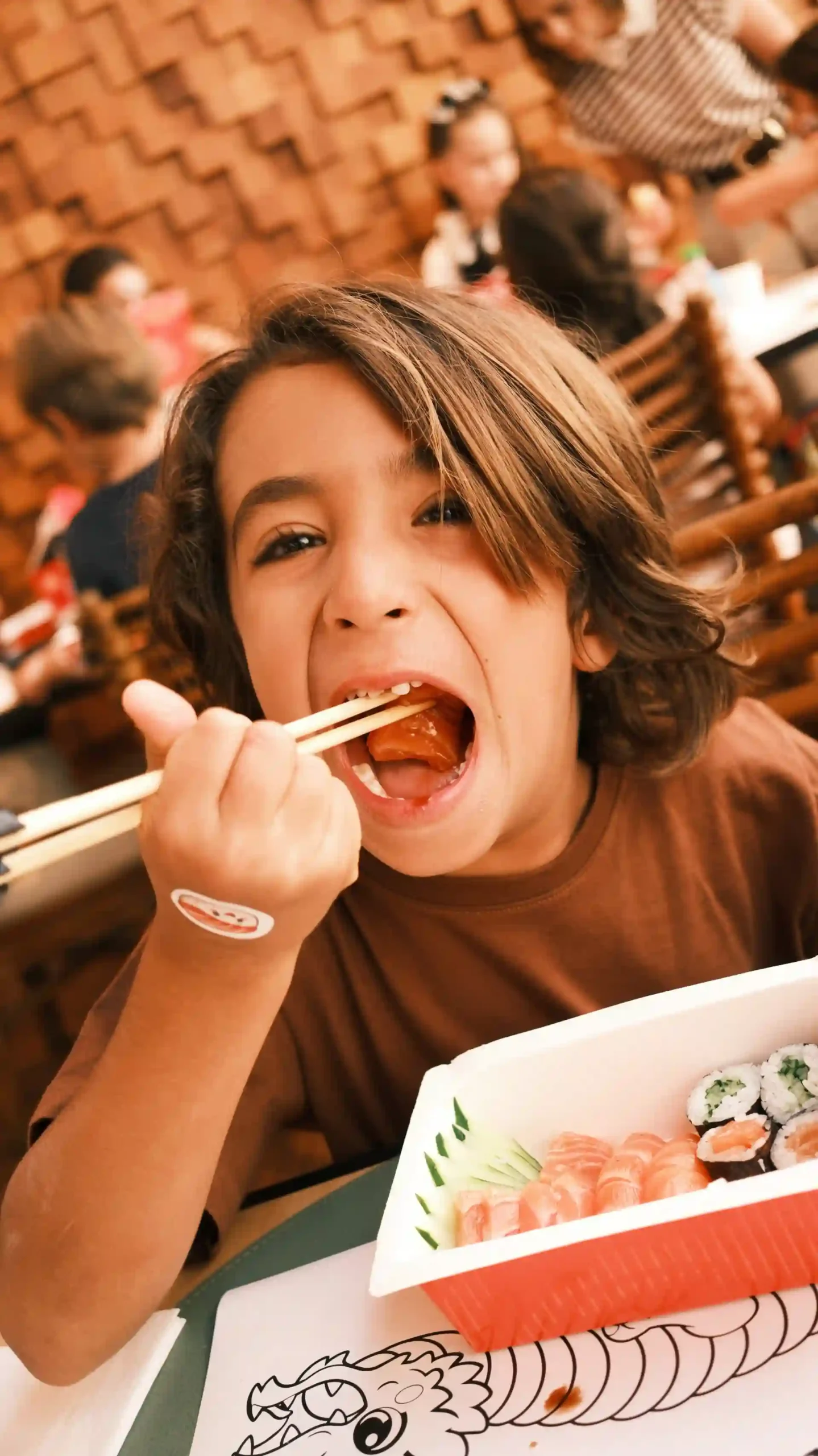 Criança sorridente com cabelo castanho escuro com cabelo meio ondulado e olhos castanhos, com pauzinhos comendo sushi em um restaurante japonês, ao fundo outras pessoas sentadas à mesa. Uma experiência de comida japonesa infantil.