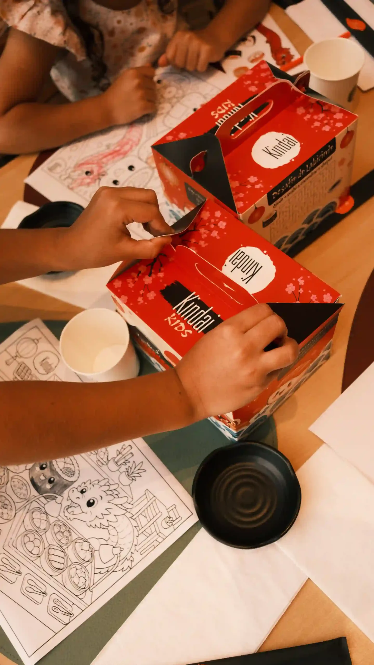 Crianças abrindo caixas de um presente de chá de boneca, com desenhos para colorir e copo de papel, em uma mesa de diversão e atividades.