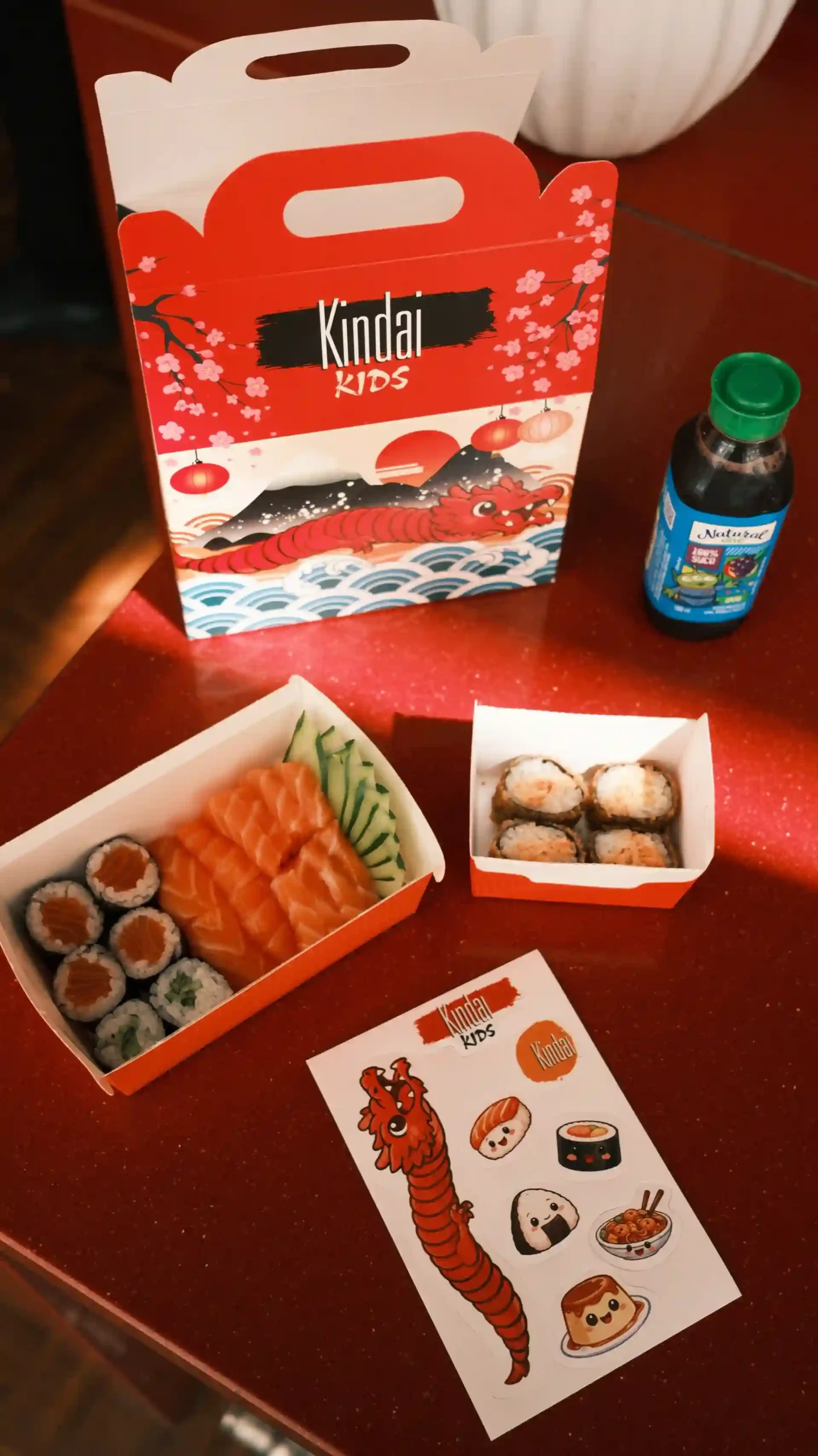 Comida japonesa incluindo salmão, sushi e chá, ao lado de um buquê de plantas verdes e uma caixa de transporte com tema de dragão para usar em festas de criança. Kindai Kids Box