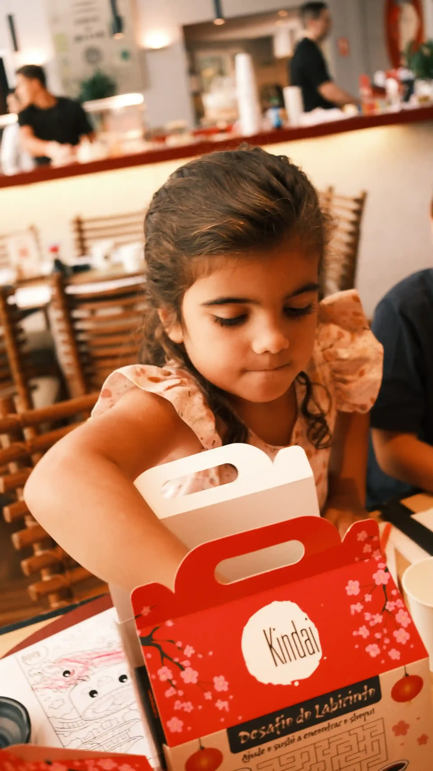 Menina colorida com cabelo castanho escuro em restaurante japonês, explorando uma caixa de comida de papel craft com rótulo "Kindai", ambiente aconchegante com decoração japonesa.