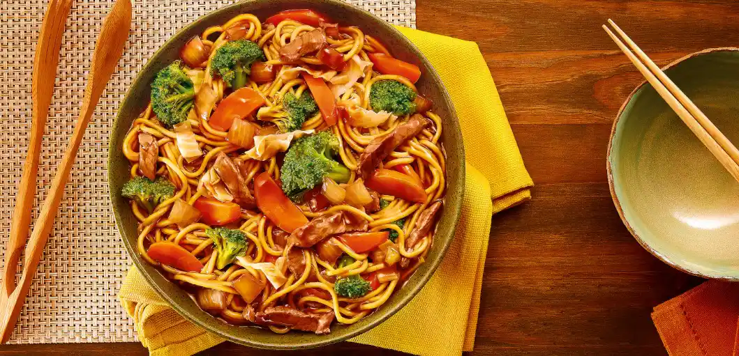 Macarrão tipo yakisoba em frigideira com legumes como brócolis, pimentão e cogumelos, acompanhado de talheres sobre mesa de madeira, pronto para servir.